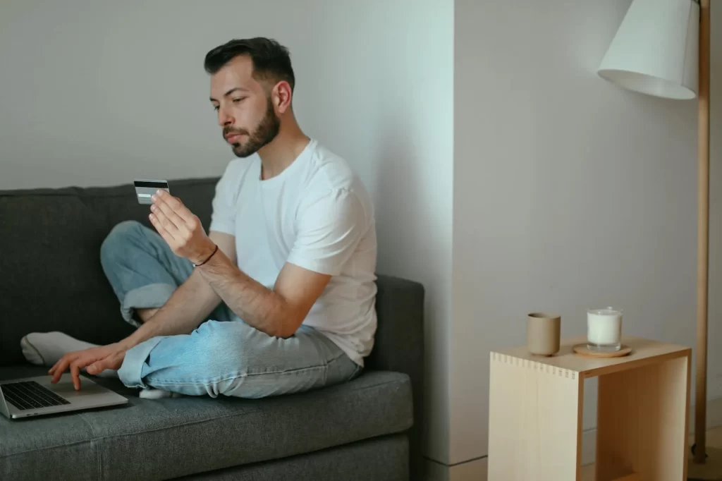 A man sitting on a couch using a laptop and holding a credit card