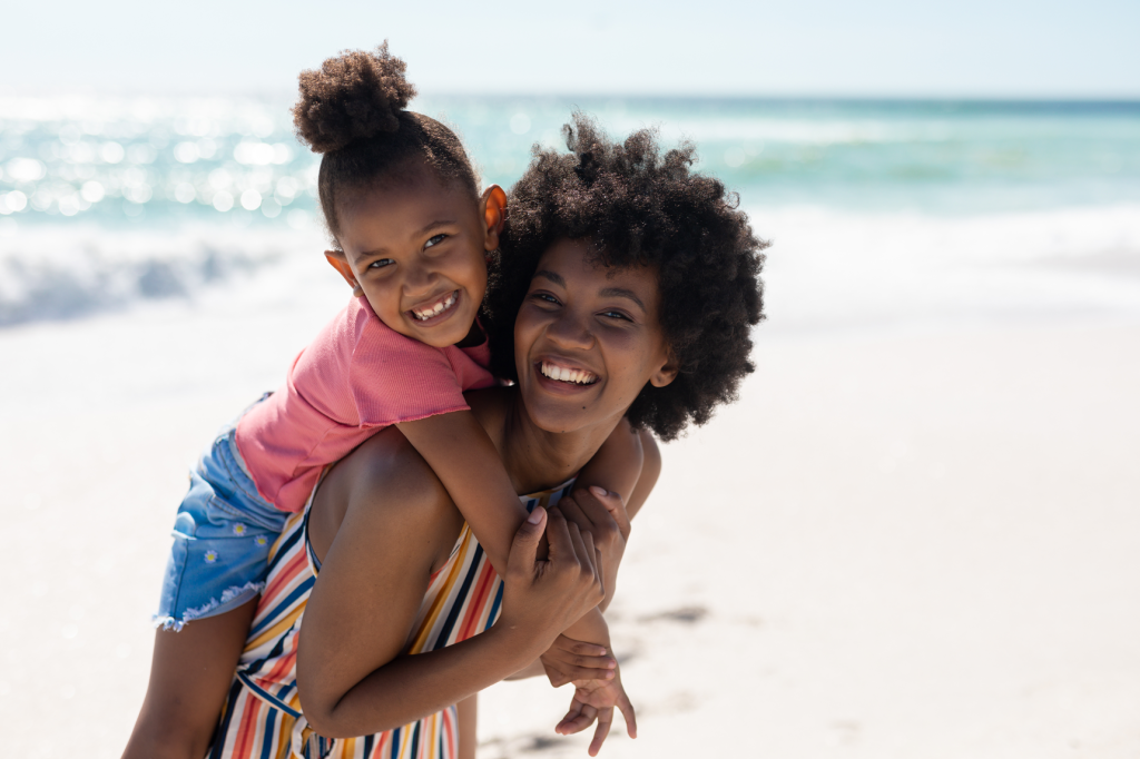A mom and daughter on the beach. They are happy and the daughter is on the mom's back. 